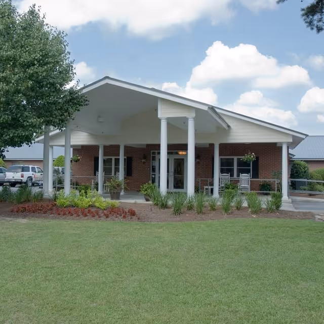 Front exterior view of a single-story brick building with a covered entrance supported by white columns. There are rocking chairs on the porch, landscaped plants and grass in the foreground, and a partly cloudy sky above.