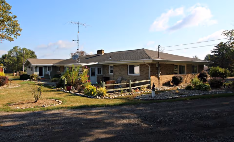 Exterior view of a single-story brick building with a sloped roof, surrounded by a garden with various plants and shrubs, under a partly cloudy sky.