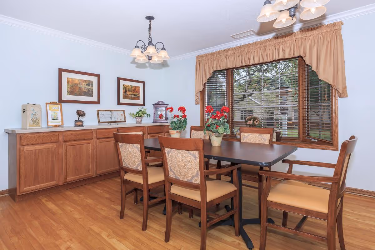 A dining room with a rectangular table surrounded by six wooden chairs with cushioned seats and backs. The table has three potted red flowering plants as decoration. Behind the table is a wooden cabinet with various decorative items and framed pictures on the wall above it. A large window with wooden blinds and a beige valance allows natural light to enter the room. The floor is wooden, and two ceiling light fixtures with multiple lampshades hang above the table.