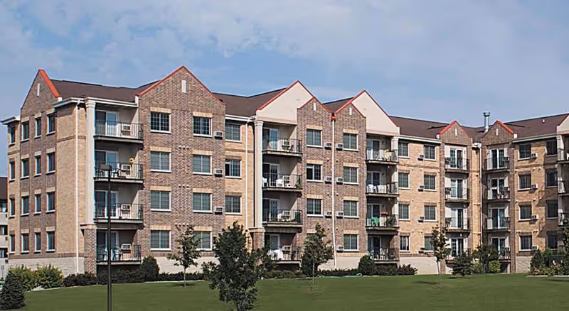 A multi-story brick senior living building with balconies, surrounded by green lawn.
