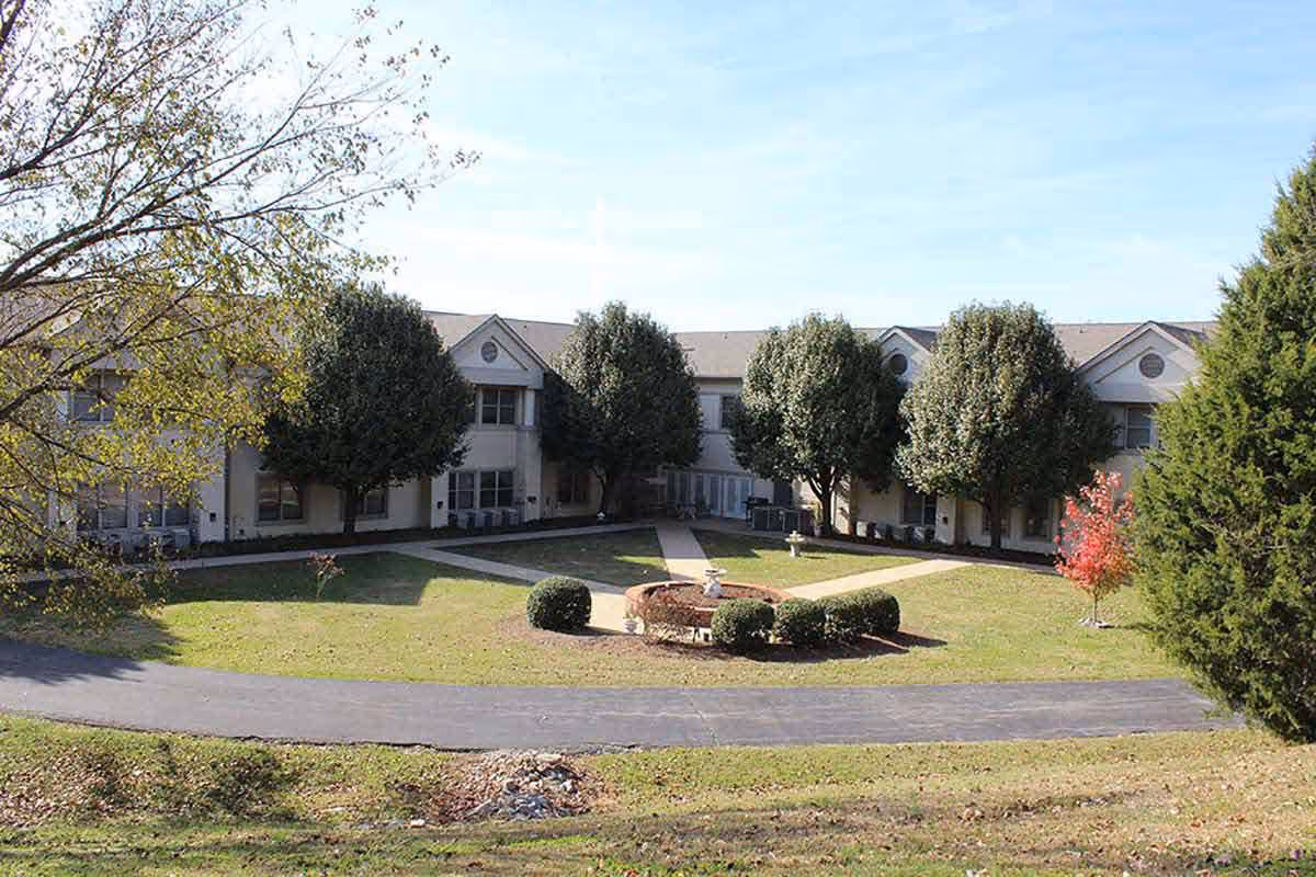 Exterior view of Poplar Estates Senior Living Community showing a two-story building surrounding a landscaped courtyard with a circular garden and trimmed bushes, several trees, and a paved walkway.
