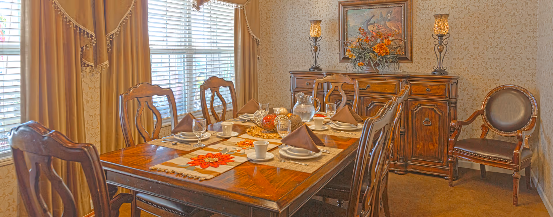A formal dining room with a wooden dining table set for six people, featuring brown napkins, white plates, cups, and glasses. The table has a decorative runner with red flower designs and a centerpiece with decorative balls. The room has beige patterned wallpaper, large windows with beige curtains, a wooden sideboard with two ornate lamps, a floral arrangement, and a framed painting of peacocks. There is also a wooden chair with a leather seat and backrest in the corner.