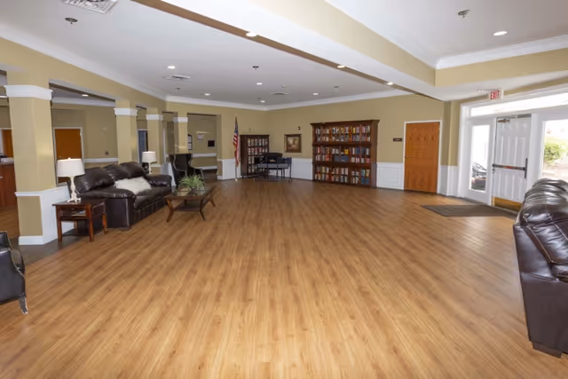 A spacious interior room with wood flooring, beige walls, and white trim. The room features two dark leather sofas, a coffee table with a plant, side tables with lamps, a bookshelf filled with books, an American flag, and a piano. There is a double glass door exit on the right side letting in natural light.