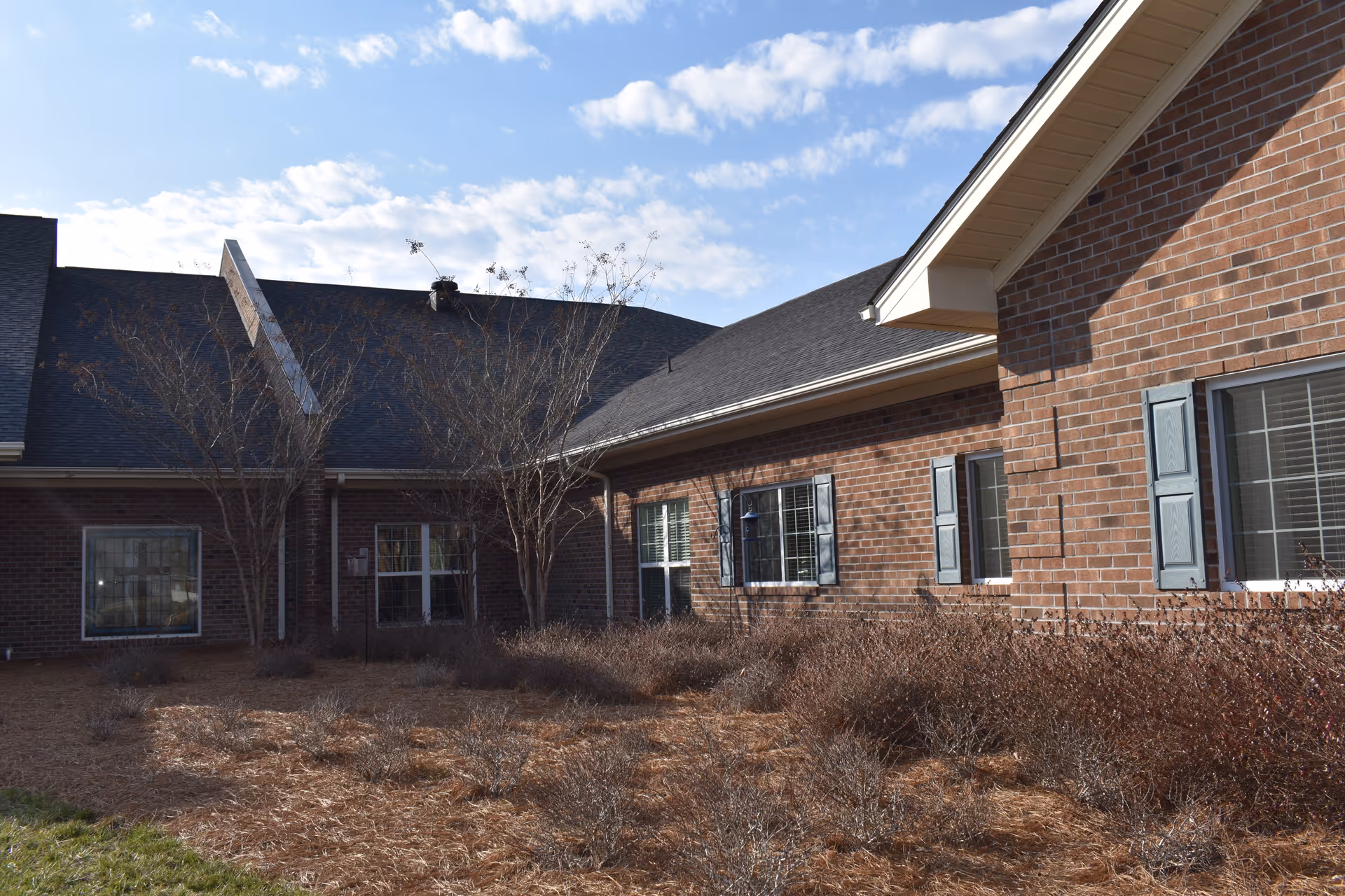 Exterior view of a brick building with multiple windows and blue shutters under a partly cloudy sky. There are leafless trees and dry bushes in front of the building.