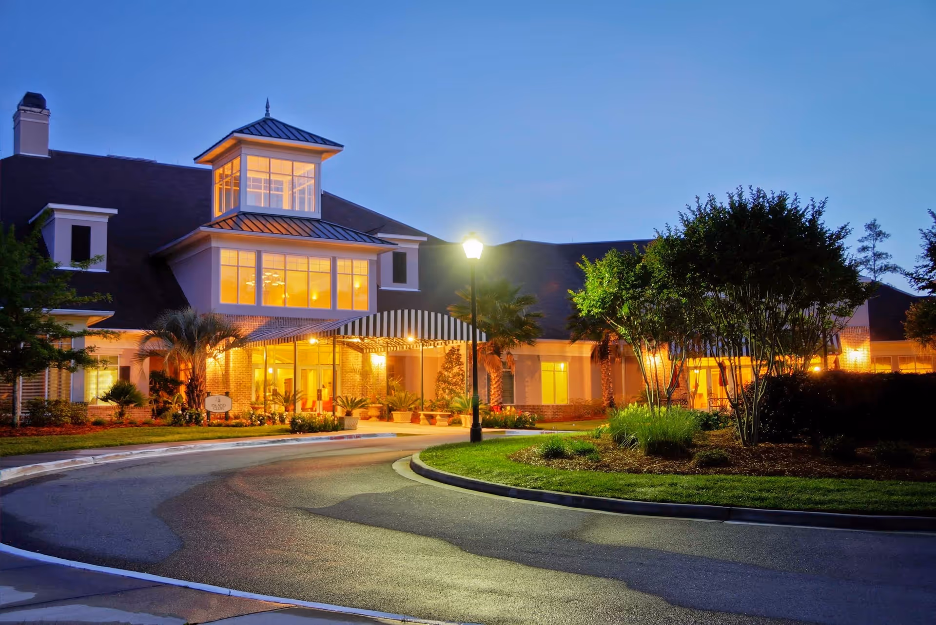 Exterior view of Marshes of Skidaway Island facility at dusk, showing a well-lit entrance with large windows, a covered driveway, landscaped greenery, and a streetlamp in front.