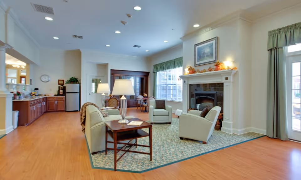A bright and spacious living room area in a senior living facility with light-colored armchairs arranged around a fireplace. The room features a patterned area rug, a wooden side table with a lamp, large windows with green curtains, and a kitchen area with wooden cabinets and a refrigerator in the background.