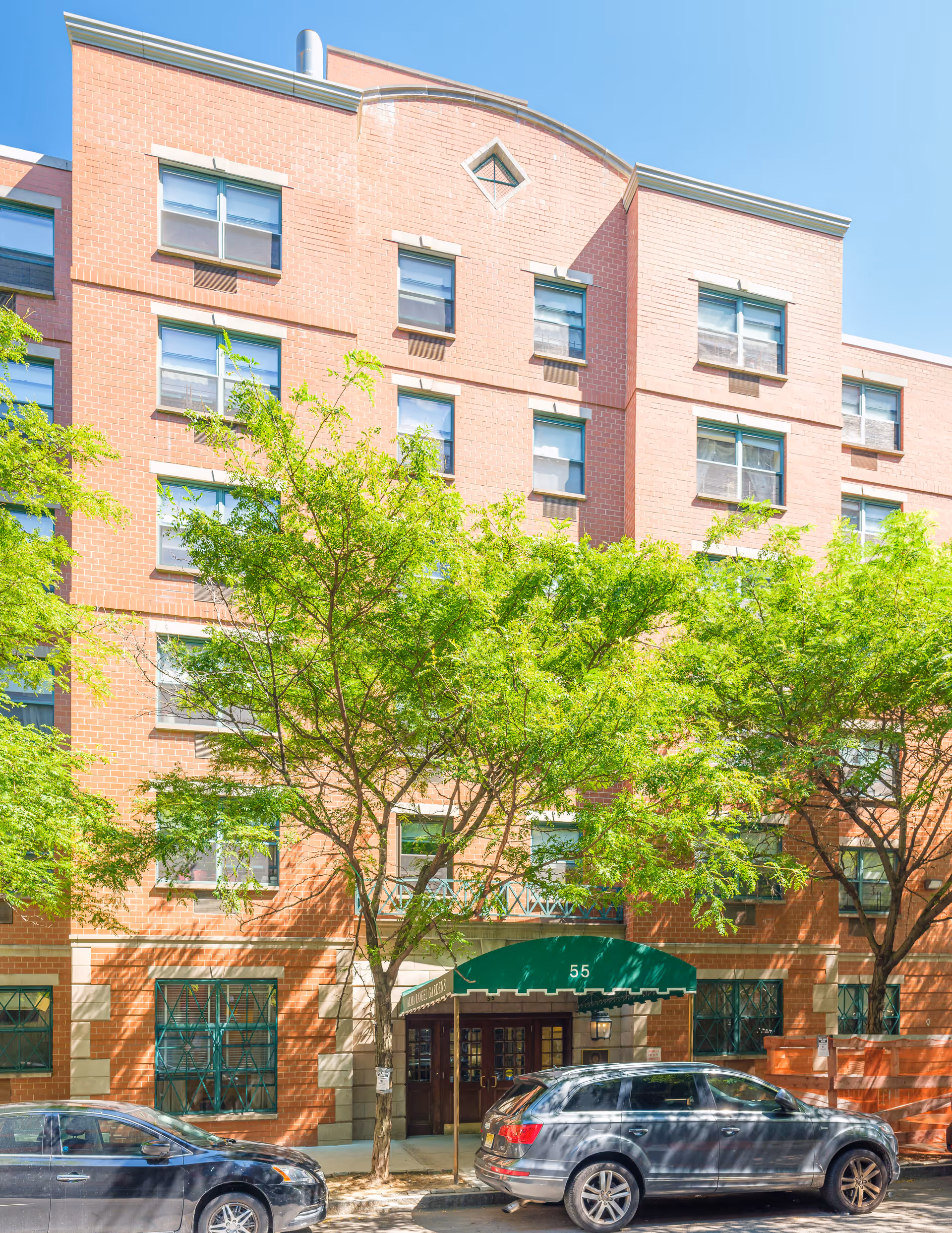 Exterior view of a multi-story brick building with several windows, green trees in front, and two parked cars along the street. The entrance has a green awning with the number 55 displayed.