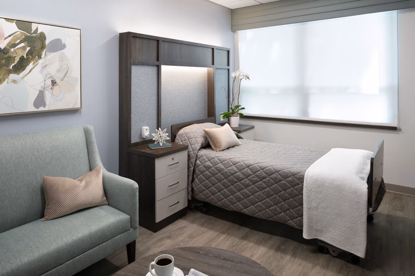 A neatly arranged bedroom in a senior living facility featuring a single bed with a gray quilted bedspread and a white blanket at the foot. Next to the bed is a dark wood headboard with built-in nightstands and soft lighting. A potted orchid sits on one nightstand. To the left, there is a light green upholstered loveseat with a beige pillow. A round wooden table with a cup of coffee and a newspaper is in the foreground. The room has a large window with a white roller shade and light-colored wood flooring.