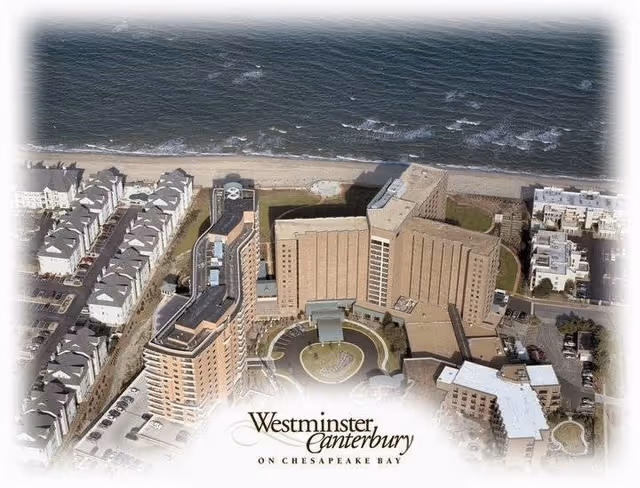 Aerial view of Westminster Canterbury on Chesapeake Bay, showing multiple large buildings near the shoreline with a beach and water in the background. The complex includes tall residential or facility buildings surrounded by parking areas and smaller structures.