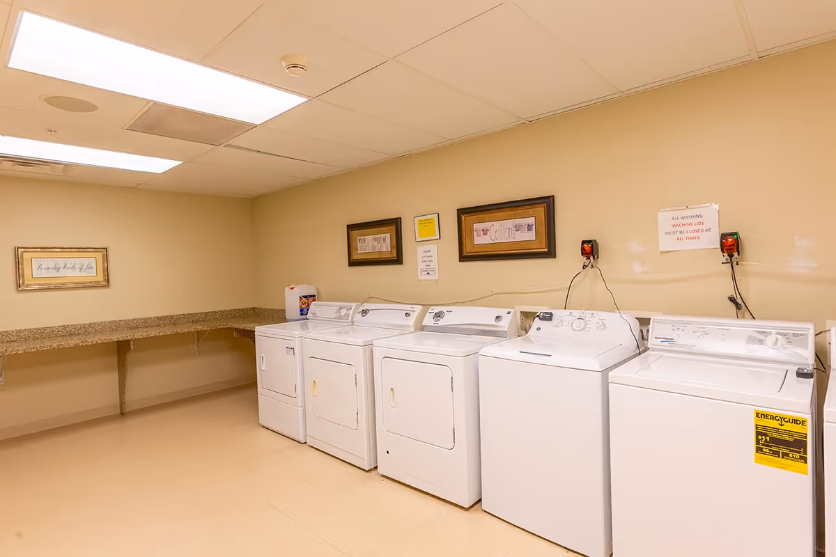Laundry room with four white washing machines and dryers lined up against a beige wall. Above the machines are framed signs labeled 'Cottons' and other laundry-related instructions. There is a long granite countertop on the left side of the room. The ceiling has fluorescent lighting panels.