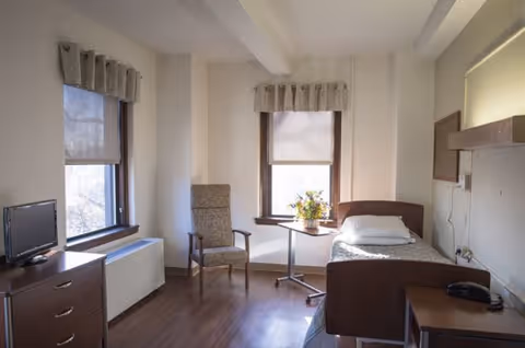 A simple, clean bedroom with a single bed, a pillow and blanket, a small bedside table with a telephone, a wooden chair, a small round table with a flower arrangement, two windows with curtains, a small TV on a dresser, and wooden flooring.