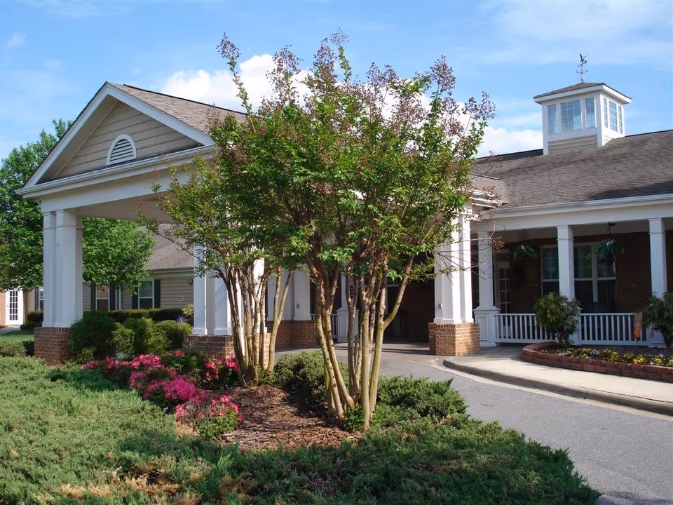 Entrance of a senior living facility with a covered driveway supported by white columns, surrounded by green shrubs, flowering plants, and trees under a blue sky.