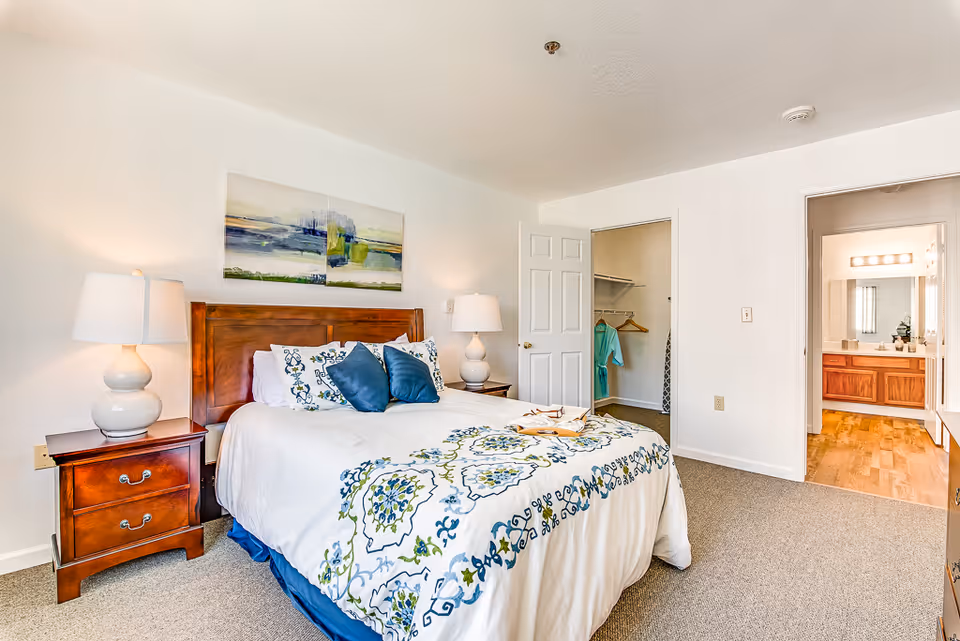 A bright bedroom with a wooden bed frame, white and patterned bedding, two bedside tables with lamps, a painting above the bed, an open closet with clothes hanging, and a view into a bathroom with a large mirror and wooden cabinets.