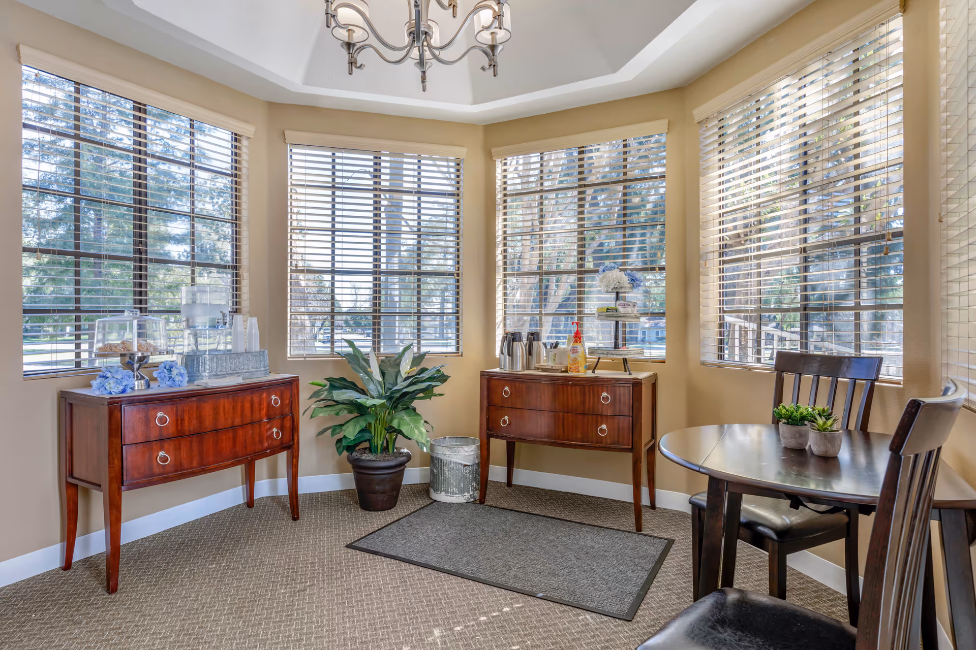 Sunny seating area with a round table and chairs, two wooden sideboards with refreshments, and large windows with blinds.