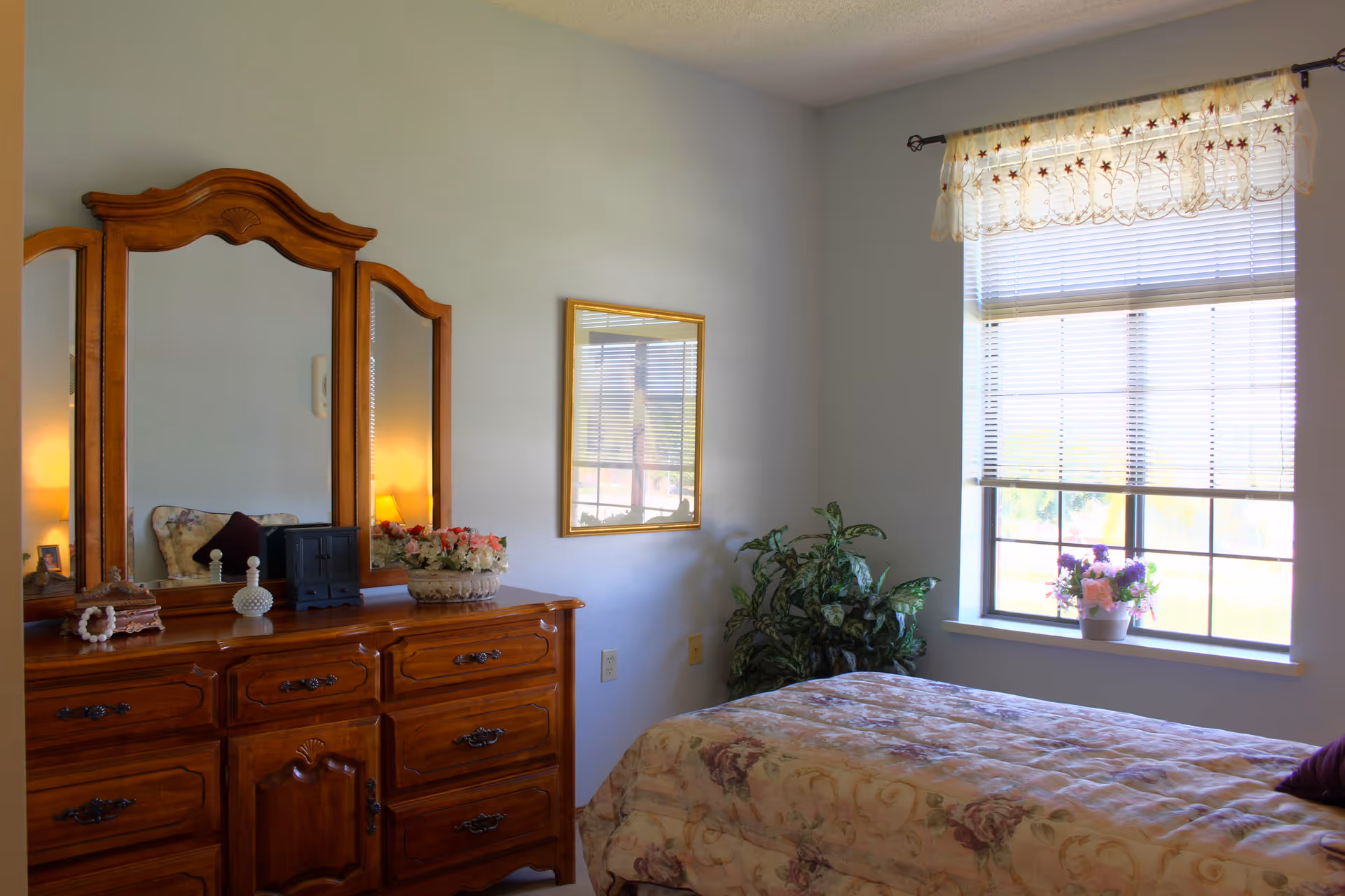 Sunlit bedroom with a wooden dresser and mirror, a bed with a floral comforter, a potted plant, and a window with blinds and a valance.