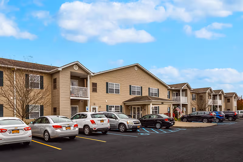 Exterior front view of a two-story beige senior living apartment building with a parking lot and cars in front.