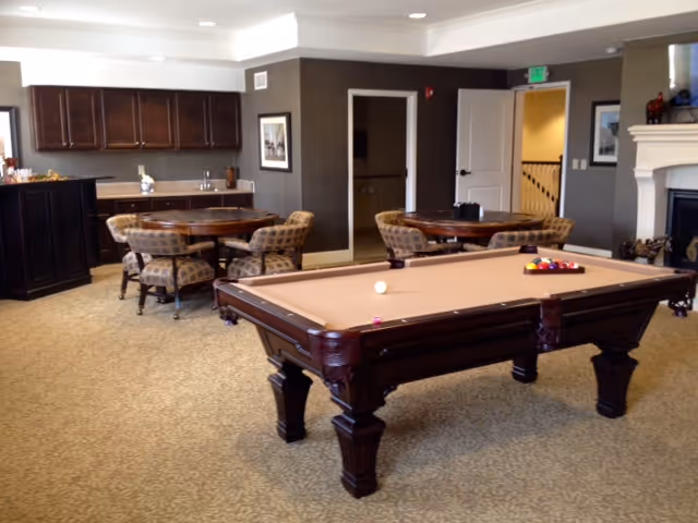 A spacious common area in a senior living facility featuring a pool table in the foreground, several round tables with cushioned chairs, dark wood cabinetry along the back wall, and a fireplace on the right side. The room has neutral-colored walls and carpeted flooring.