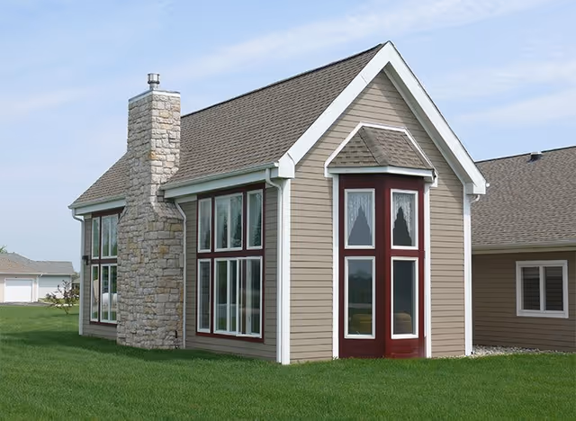 Exterior view of a beige house with a stone chimney, large windows, and a steep roof, surrounded by green grass under a partly cloudy sky.