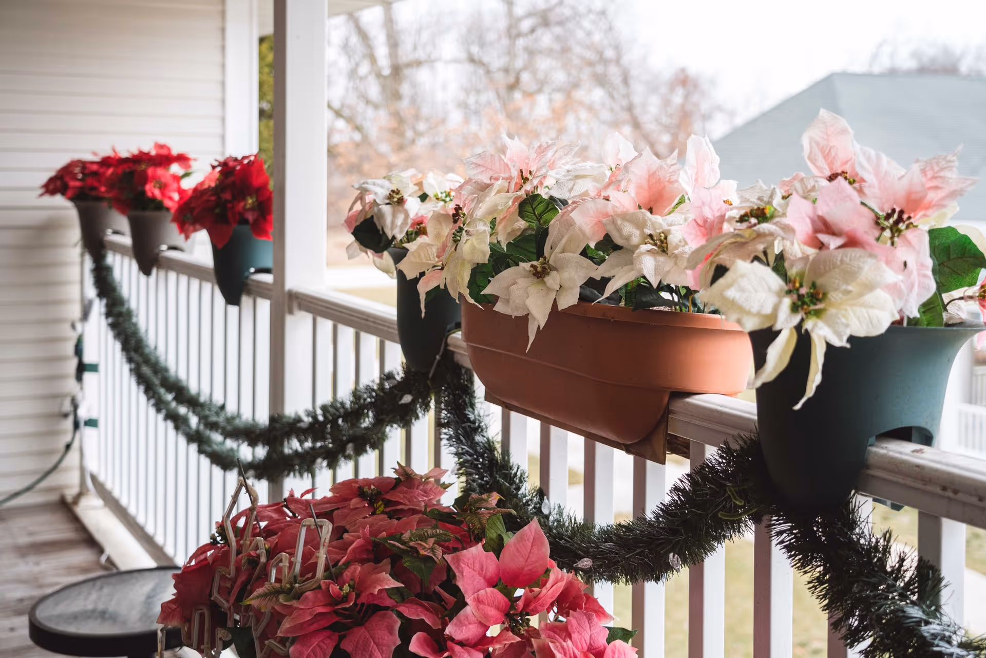A porch railing decorated with garland and multiple pots of poinsettia plants in red, white, and pink colors. The background shows a blurred view of trees and rooftops.