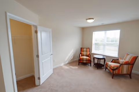 Carpeted sitting room with two striped armchairs and a round side table by a large window and an open closet door.