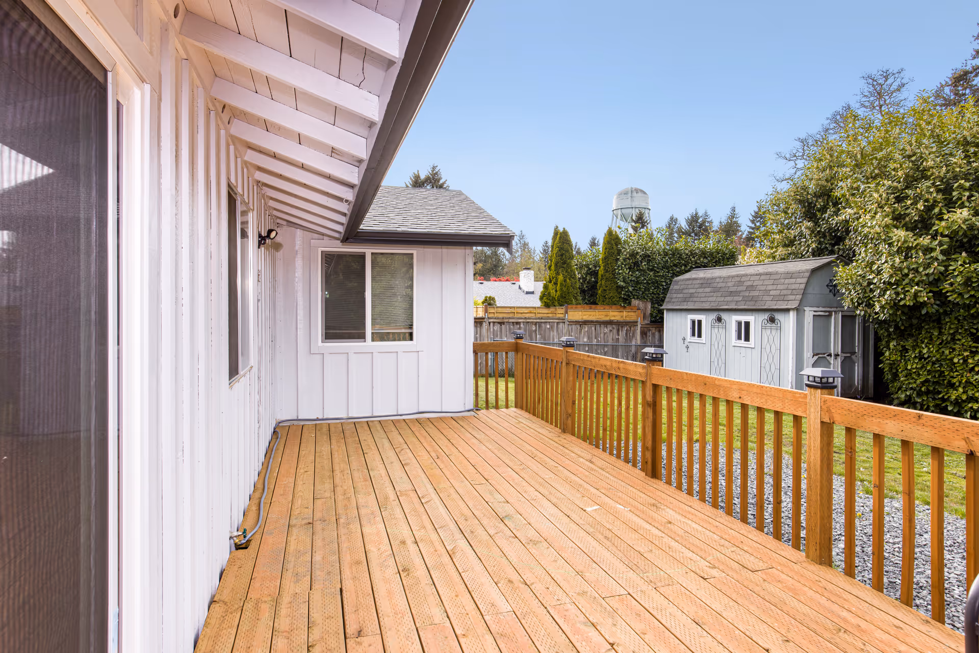 A wooden deck attached to a white building with a slanted roof. The deck has a wooden railing with small lantern-style lights on the posts. In the background, there is a small gray shed, a fenced yard with green grass, trees, and a water tower visible in the distance under a clear blue sky.