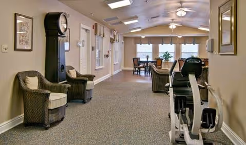 Interior view of a senior living facility hallway with beige walls and carpeted floor. On the left side, there are two wicker chairs with cushions and a tall grandfather clock. Further down the hallway, there is a seating area with tables and chairs near large windows with blinds. On the right side, there is an exercise bike and a mirror on the wall. The ceiling has recessed lighting and ceiling fans.