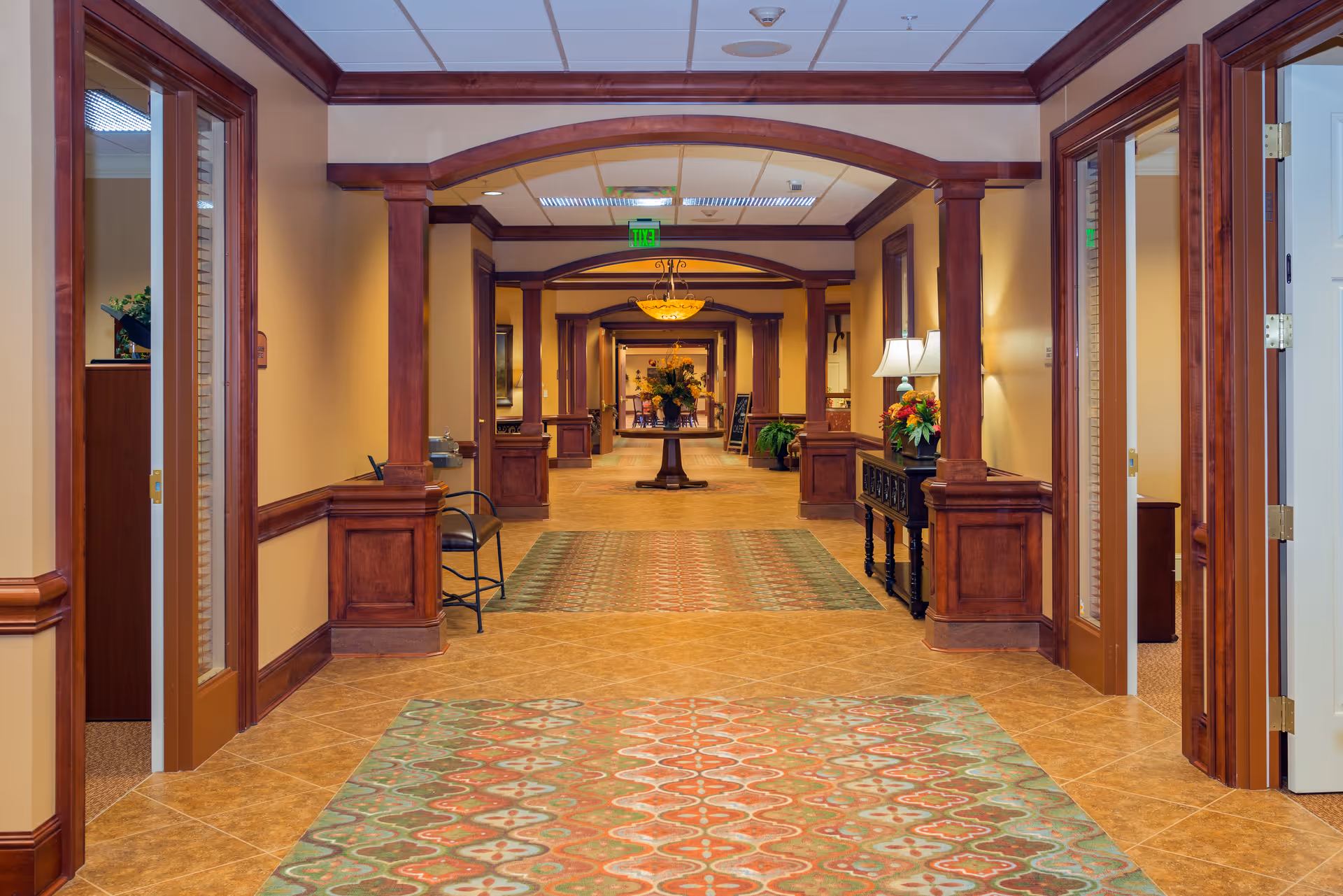 A long, well-lit hallway in a senior living facility with wooden archways and trim, patterned rugs on the tiled floor, chairs along the walls, and decorative plants and lamps on side tables.