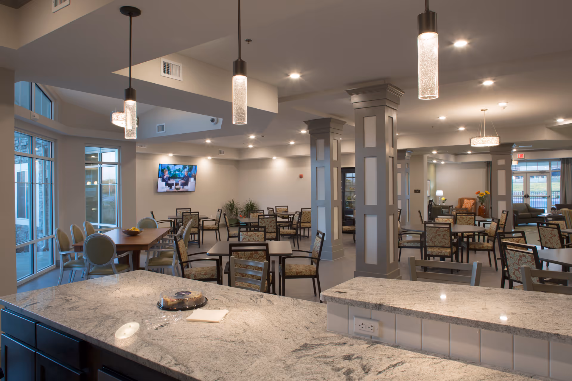 Spacious communal dining and lounge area with multiple tables and chairs, pendant lights, and a granite countertop in the foreground.