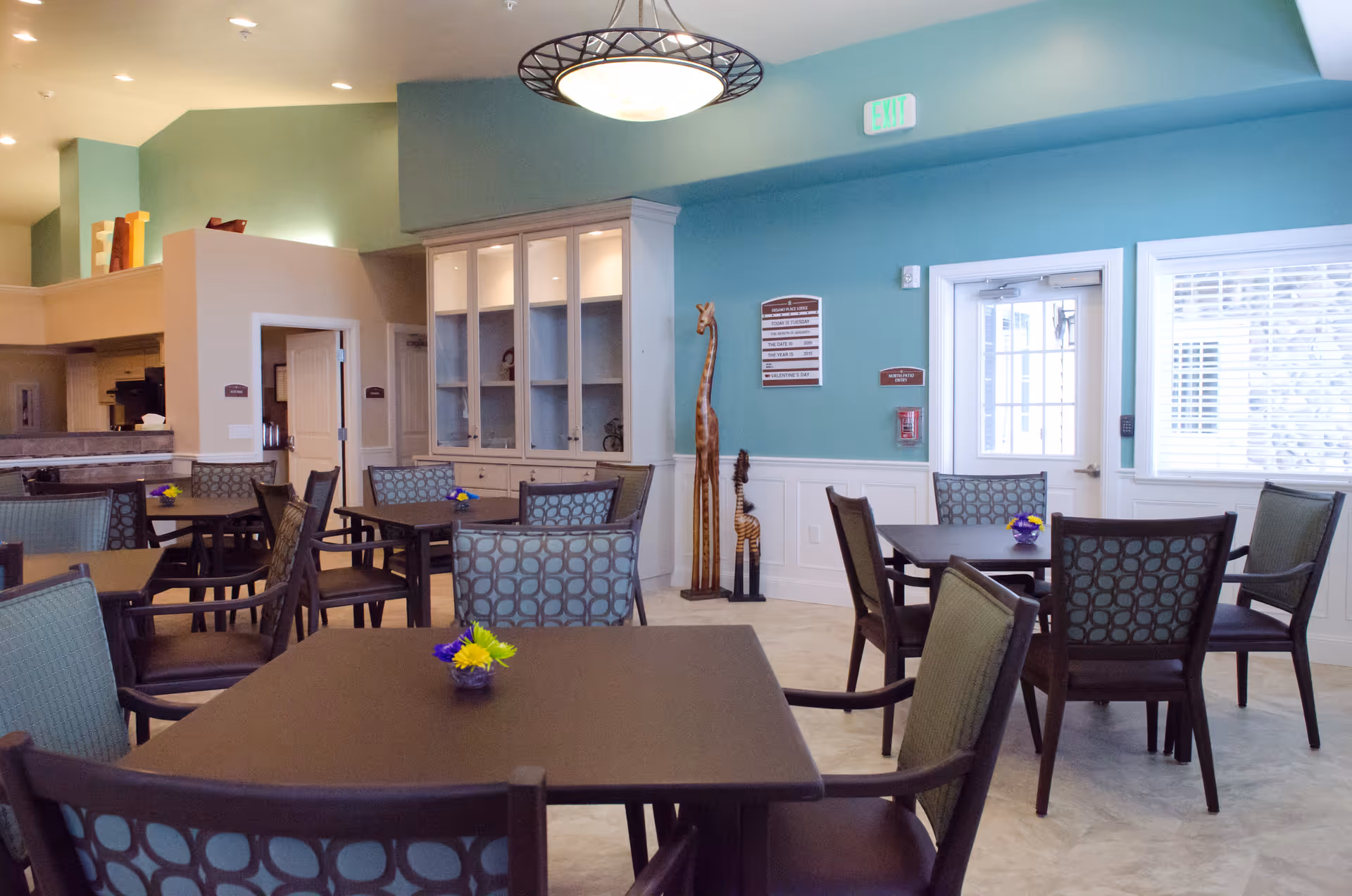 A bright and clean dining area with multiple square tables and cushioned chairs arranged neatly. The walls are painted teal with white wainscoting, and there is a white cabinet with glass doors against the wall. Two tall wooden giraffe sculptures stand near the cabinet. A door with glass panels and a window with blinds are visible on the right side. Small flower arrangements are placed on each table.