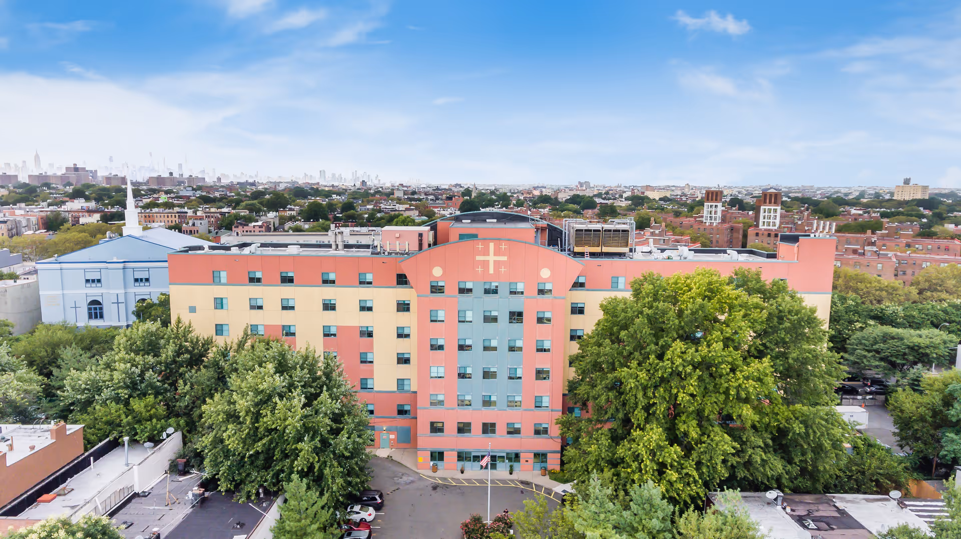 Aerial view of a multistory, multicolored nursing facility building front surrounded by trees with a city skyline in the background.