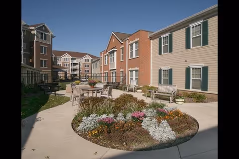 Outdoor courtyard area at Oak Park Place Madison featuring a circular flower bed with colorful flowers in the center, surrounded by a paved walkway. There are several benches and tables with chairs arranged around the courtyard. The courtyard is bordered by multi-story residential buildings with brick and siding exteriors under a clear blue sky.