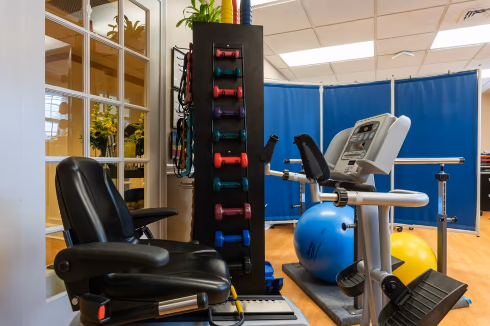 Small exercise/therapy room with a recumbent exercise machine, dumbbell rack, exercise balls and a black chair.