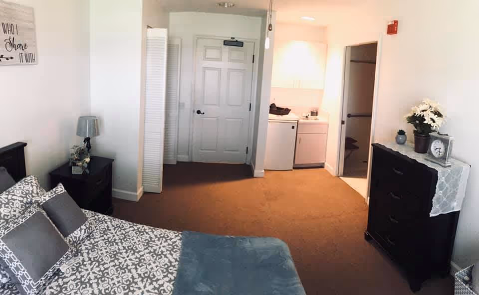 A bedroom with a patterned bedspread and multiple pillows on the bed. There is a nightstand with a lamp and decorative items next to the bed. Across the room, there is a dresser with a lace runner, a clock, a small plant, and a vase with white flowers. The room has a door leading to a small kitchenette area with a mini fridge and sink, and another door leading to a bathroom or closet. The walls are white, and the carpet is brown.