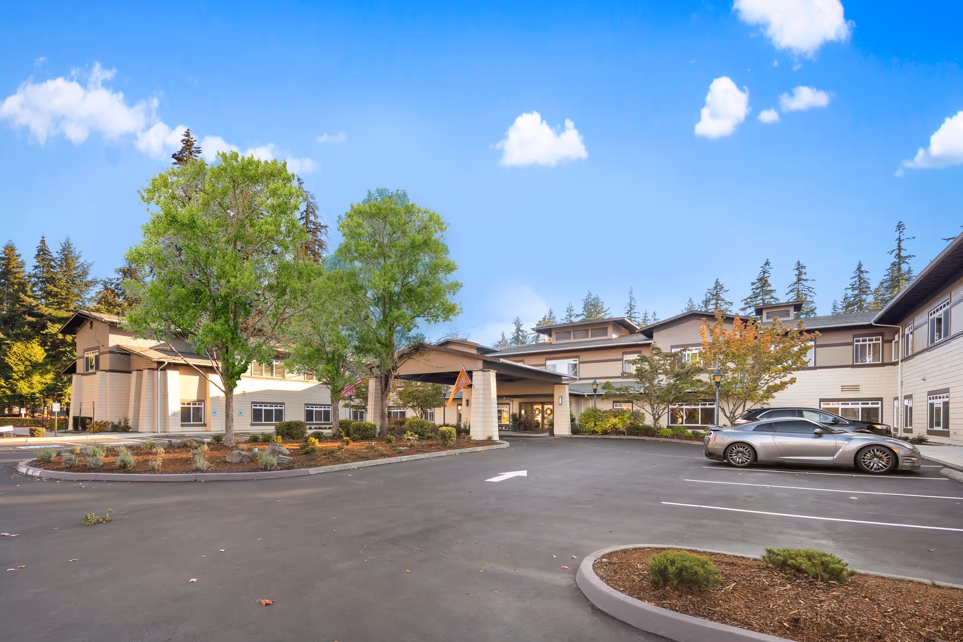 Exterior view of Sunrise of Redmond senior living facility showing a two-story building with a covered entrance, surrounded by trees and a parking lot with a silver car parked.