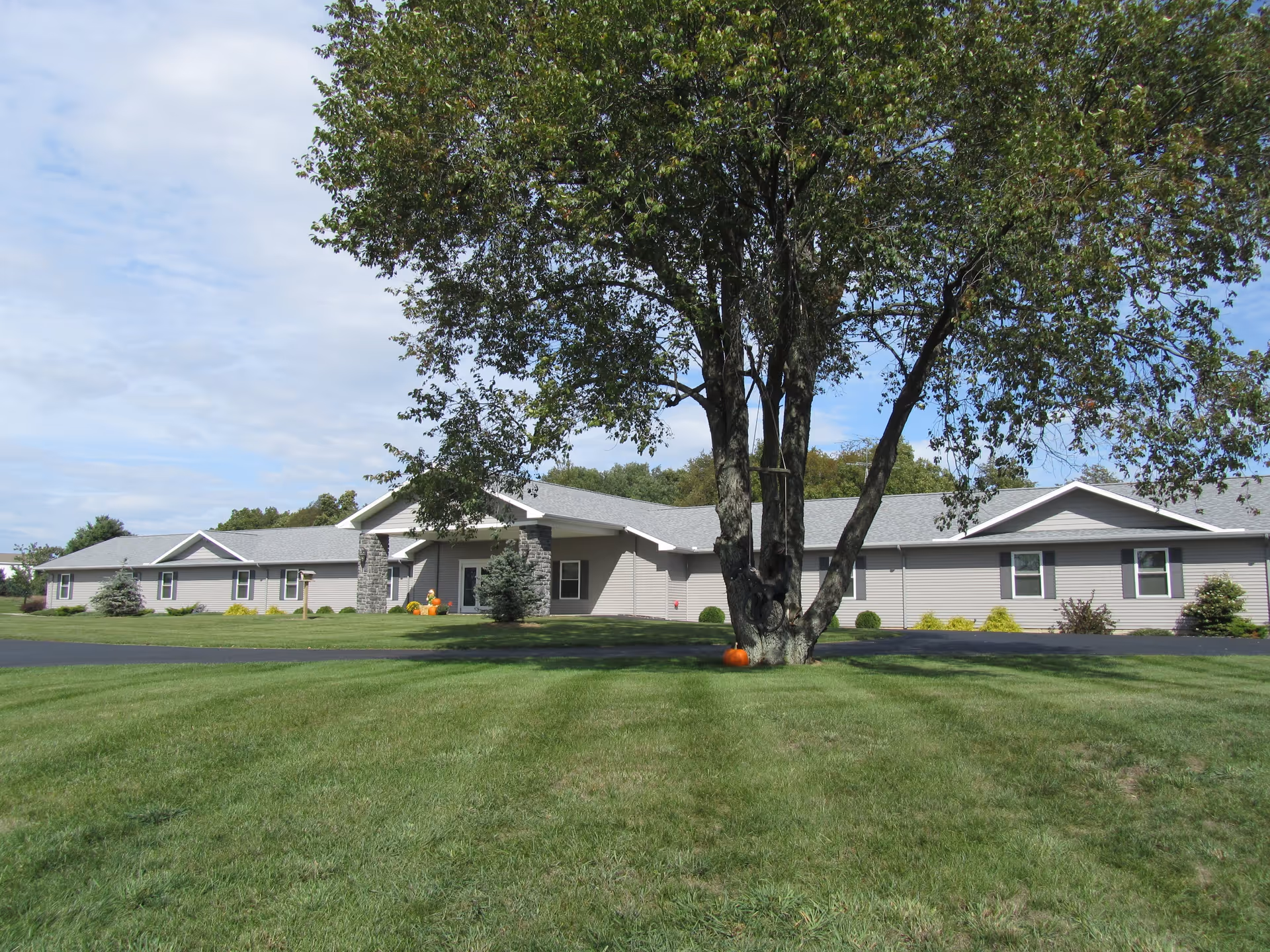 A single-story building with a gray roof and beige siding, surrounded by a well-maintained green lawn and a large tree in the foreground. The sky is partly cloudy.