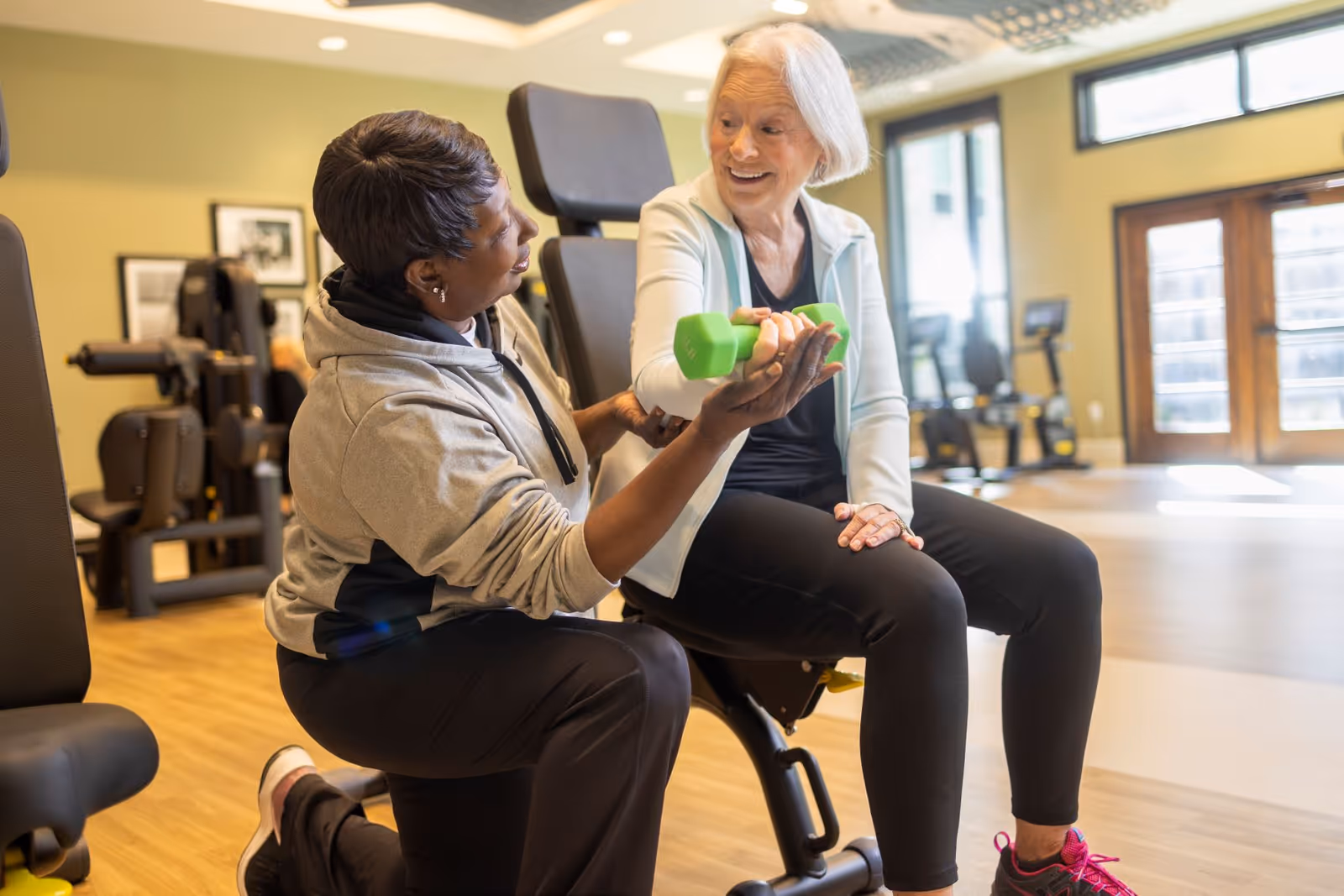 An elderly woman sitting on a workout bench lifting a green dumbbell with the assistance of a fitness trainer in a well-lit gym room with exercise equipment in the background.