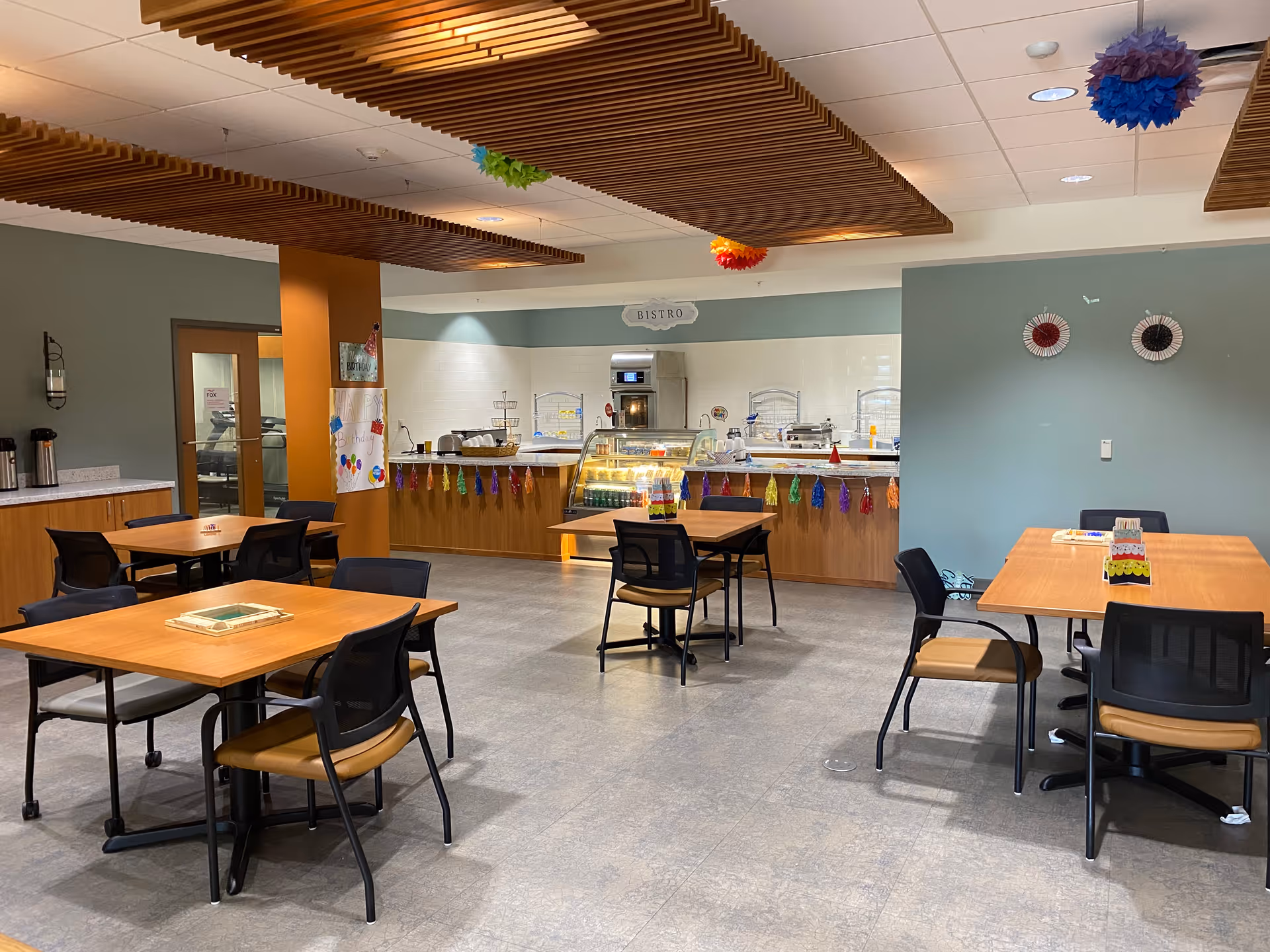 A dining area in an assisted living facility with several wooden tables and black chairs arranged neatly. The room features a serving counter labeled 'Bistro' with food display cases and colorful decorations hanging from the ceiling and counter. The walls are painted in muted tones with some decorative wall hangings, and the ceiling has wooden slats adding texture to the space.