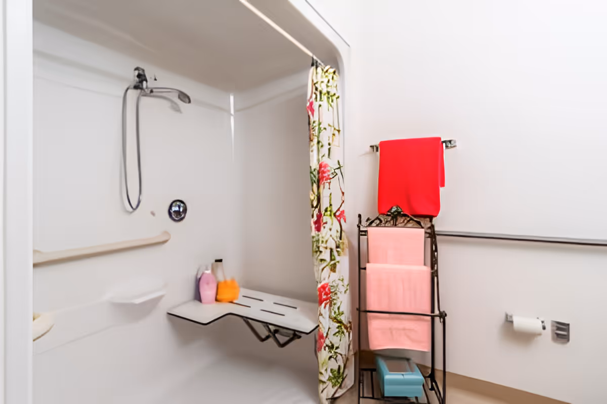 A white accessible shower area with a fold-down seat, a handheld showerhead, and grab bars. There is a floral shower curtain partially drawn to the side. Next to the shower, there is a metal towel rack holding pink and red towels, and a toilet paper holder mounted on the wall.