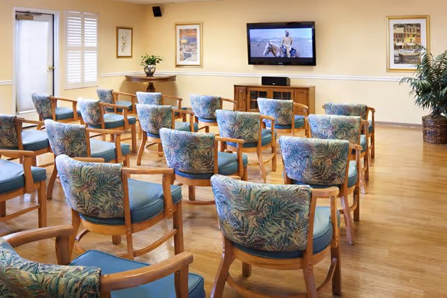 A senior living facility common room with multiple wooden chairs arranged in rows facing a wall-mounted television. The chairs have blue cushions and patterned backs. The room has light yellow walls, wooden flooring, framed artwork, a potted plant, and a door with window blinds.