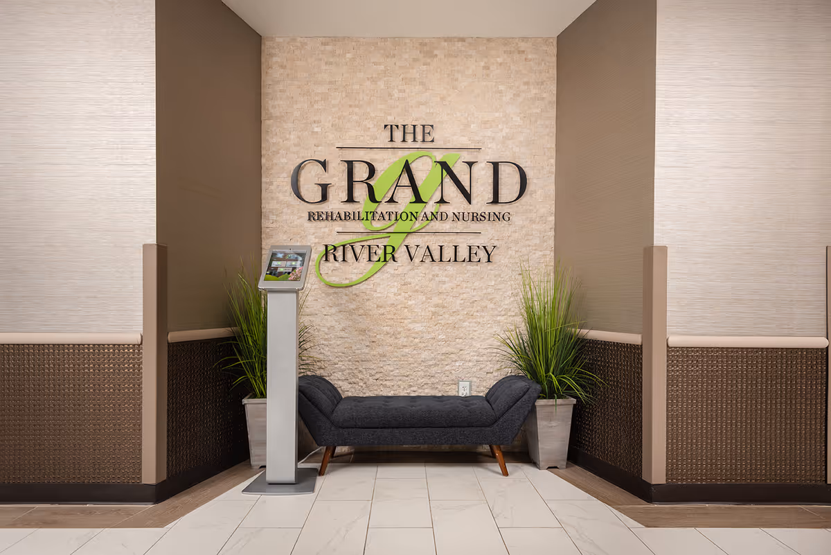 Interior view of a reception or waiting area at The Grand Rehabilitation and Nursing at River Valley, featuring a textured stone wall with the facility's name displayed, a dark gray cushioned bench, two potted plants on either side, and a digital kiosk stand.