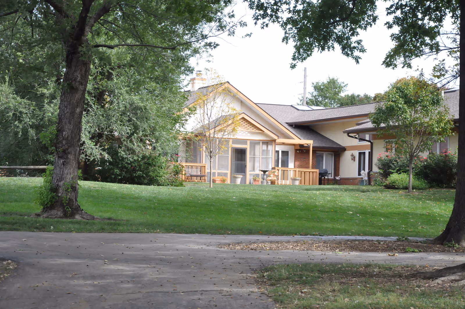 Single-story residential building with a screened porch and wooden deck set behind a grassy lawn and trees.