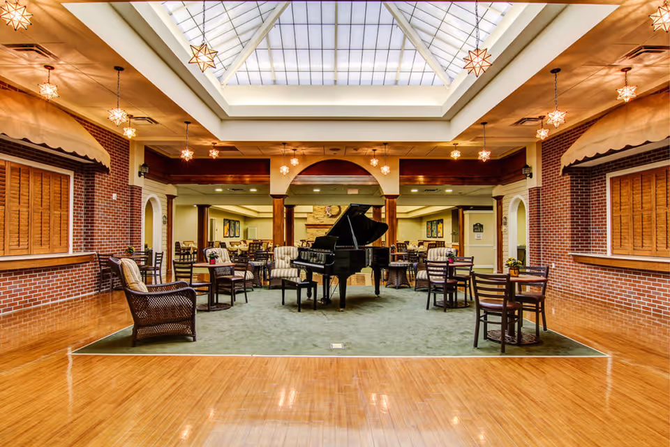 Spacious senior living common area with a grand piano under a large skylight, surrounded by tables, chairs, and wicker seating.