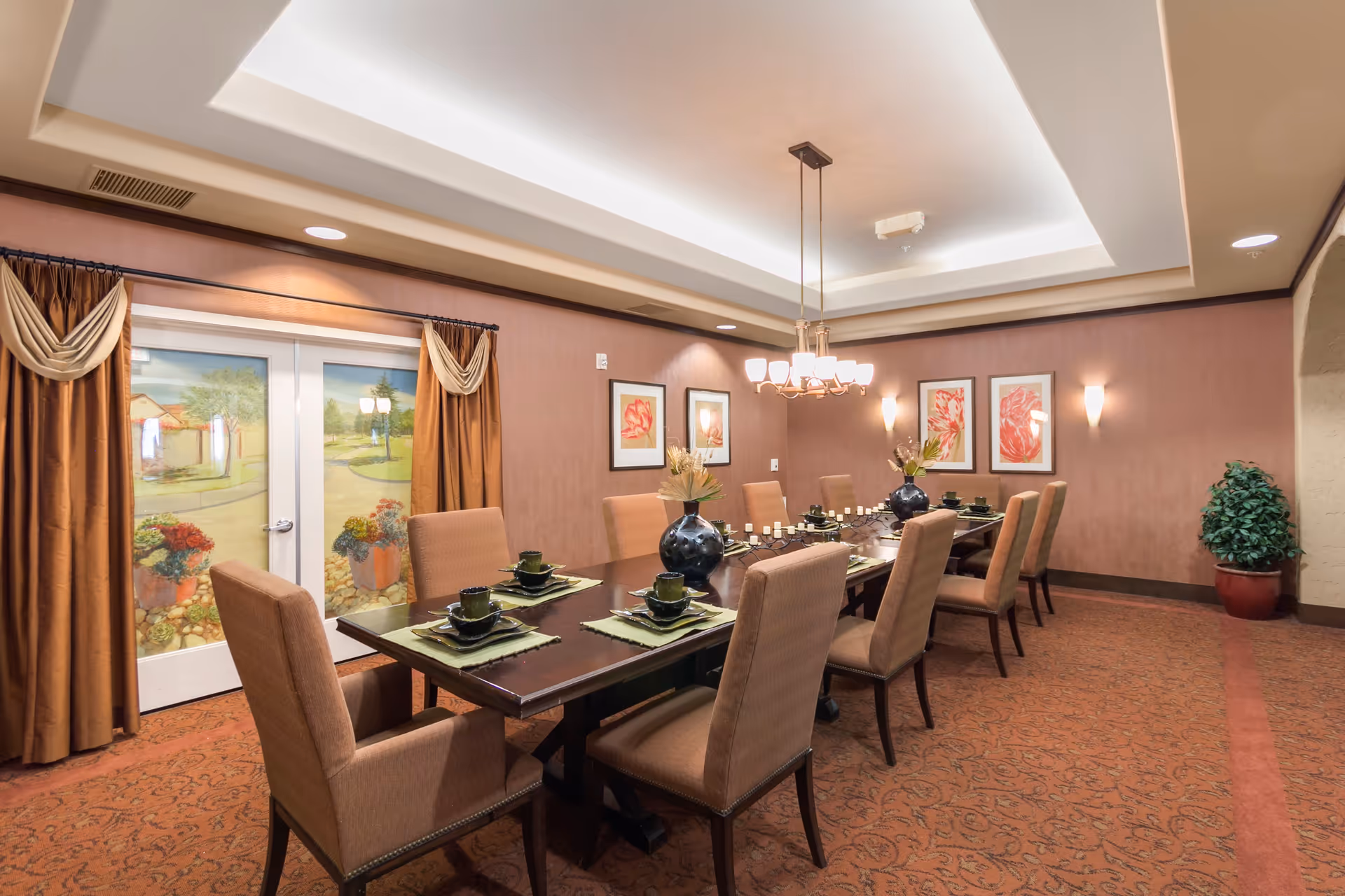 A formal dining room with a long dark wooden table set for eight people. The table has green placemats, black plates, cups, and decorative black vases with flowers. The room features brown upholstered chairs, warm brown walls with framed floral artwork, a chandelier hanging from a recessed ceiling, and double doors with painted glass panels depicting an outdoor scene. There is a potted plant in the corner and brown curtains framing the doors.
