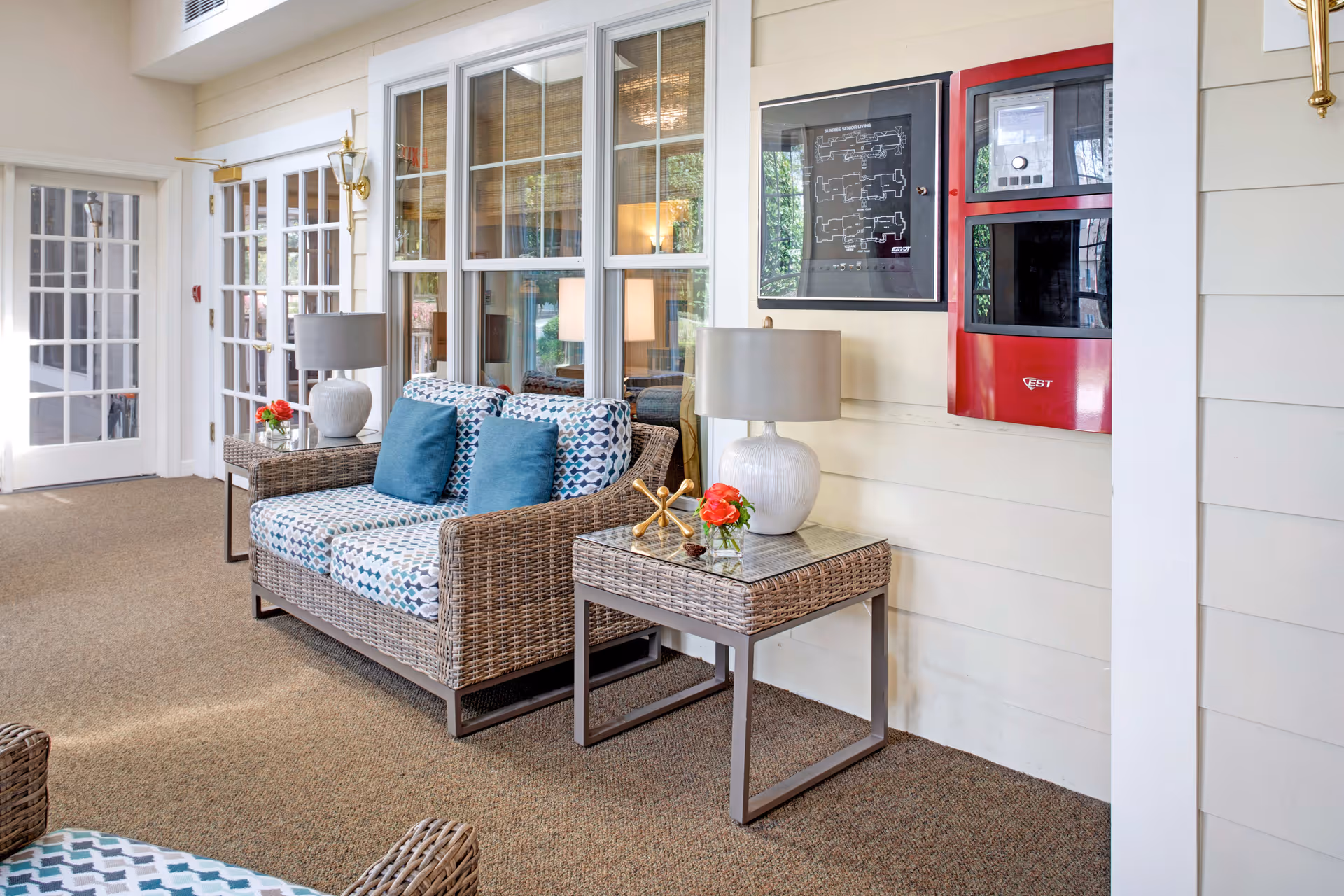 A cozy seating area with a wicker loveseat featuring patterned cushions and blue throw pillows, accompanied by a wicker side table with a white lamp and a small vase with red flowers. The area has beige carpet flooring, white paneled walls, and large windows with white frames. A red emergency panel and a framed floor plan are mounted on the wall. French doors with glass panes are visible in the background.