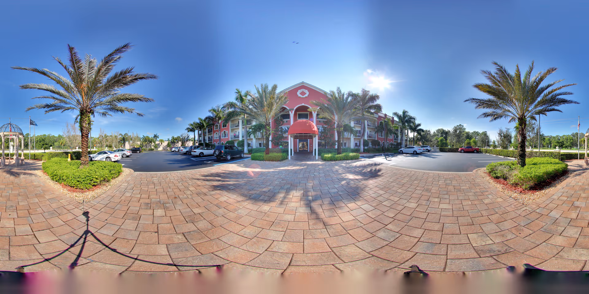 Wide panoramic view of the front exterior of Tuscany Villa of Naples, showing a three-story building with a red facade and white trim. The entrance has a red awning and is flanked by palm trees and landscaped greenery. There is a paved driveway and parking area with several parked cars under a clear blue sky.