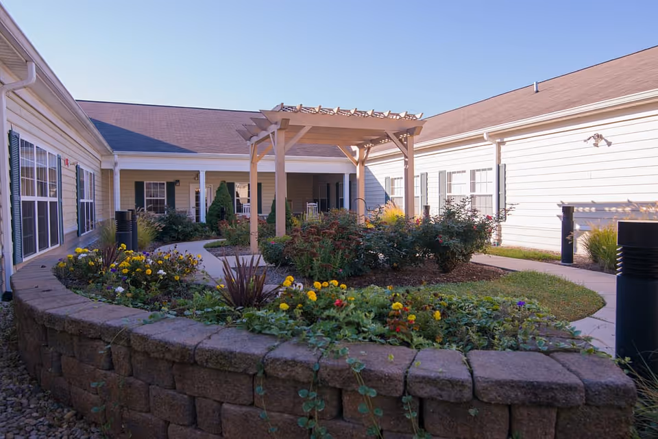 Outdoor courtyard garden area at Morning Pointe of Franklin with a wooden pergola, flower beds, shrubs, and a stone retaining wall surrounded by a single-story building with white siding and multiple windows.