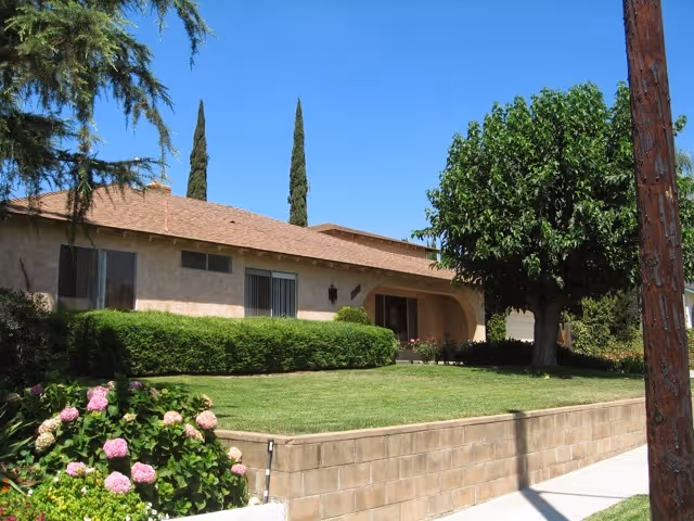 Single-story residential building with a brown tiled roof, beige exterior walls, and several windows. The front yard features a well-maintained green lawn, bushes, flowering plants, and a large tree. The sky is clear and blue.