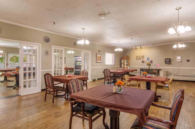 Dining room with multiple tables and chairs covered with tablecloths and small flower centerpieces in a senior living facility.