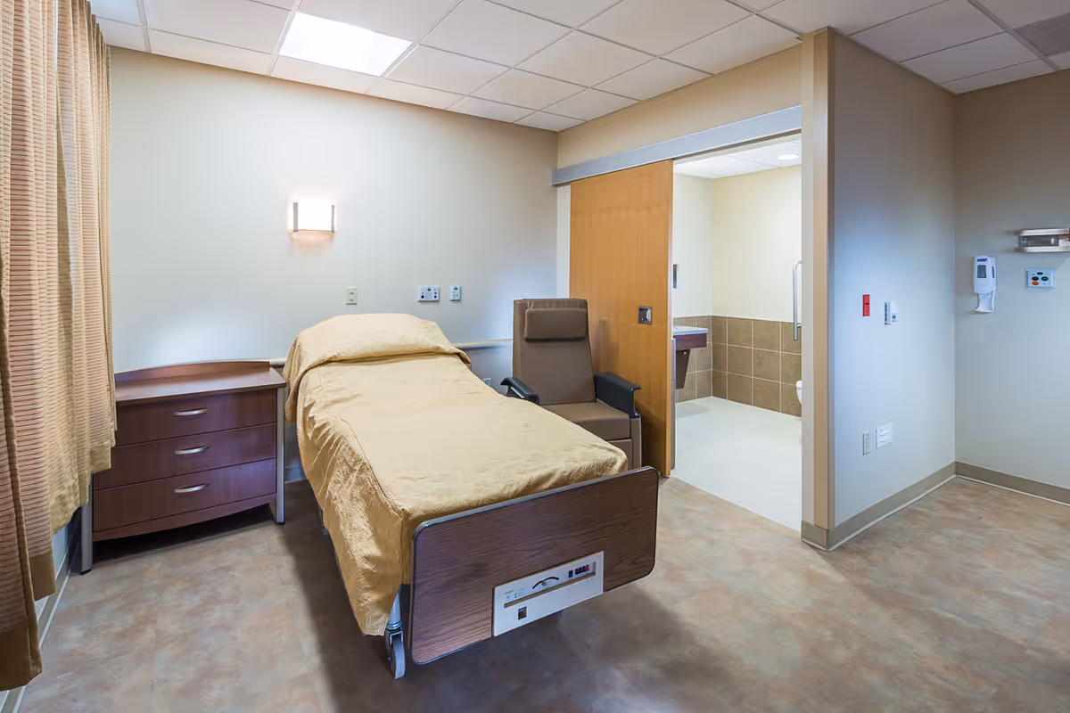 A clean and simple patient room with a single bed covered in a beige blanket, a wooden nightstand with three drawers, a brown reclining chair, and an open doorway leading to a bathroom with tiled walls and a sink.
