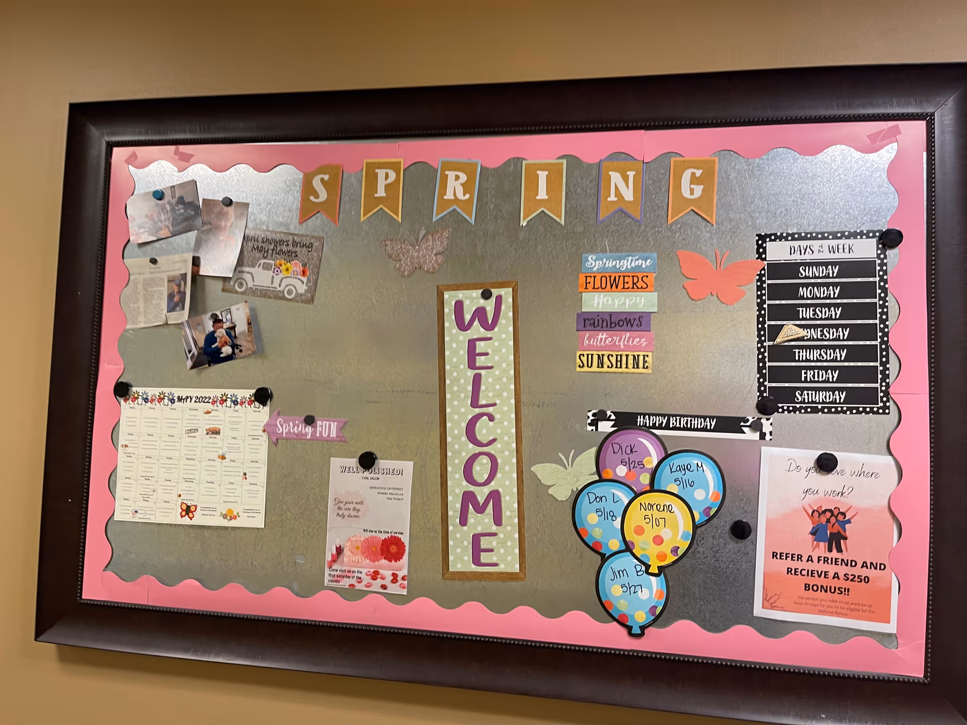 A bulletin board with a pink scalloped border and a metallic silver background. The top has a banner spelling out SPRING. The center has a vertical sign that says WELCOME. Various notes, photos, and decorations are pinned around the board, including a calendar for May 2022, a days of the week chart, birthday balloons with names and dates, and a flyer about a referral bonus. There are also spring-themed decorations like butterflies and flowers.